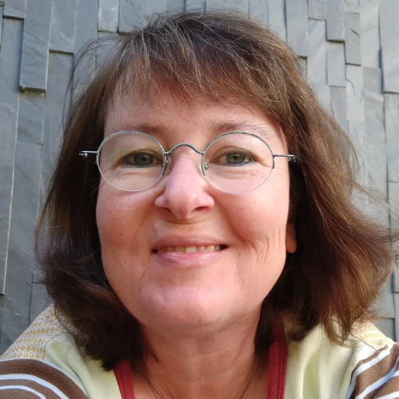 Femme brune souriante avec lunettes devant un mur avec ardoises grises